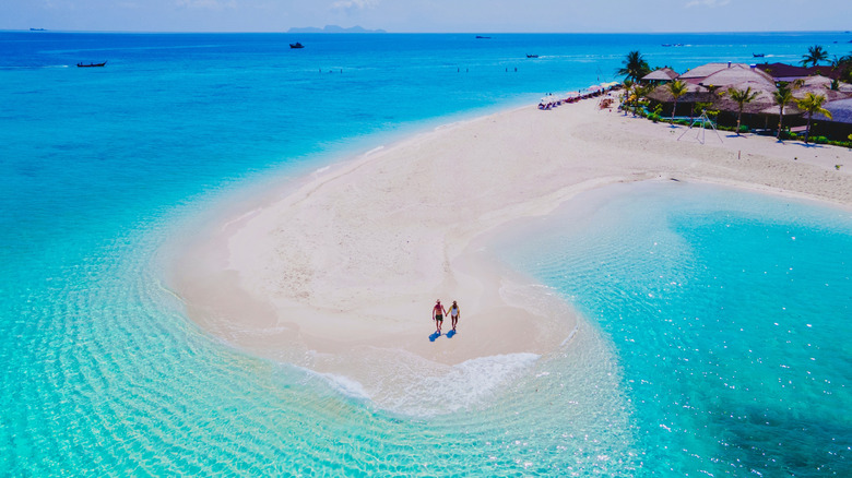A couple on a sandbar surrounded by turquoise waters in Koh Lipe, with beach huts and palm trees in the distance
