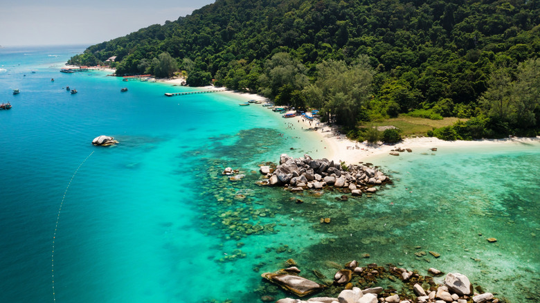 Clear blue green waters and white sand beaches backed by lush mountains with rocky shores in the foreground