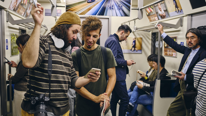 Two foreign tourists on a subway train in Japan, holding a camera and luggage, and looking at a mobile phone