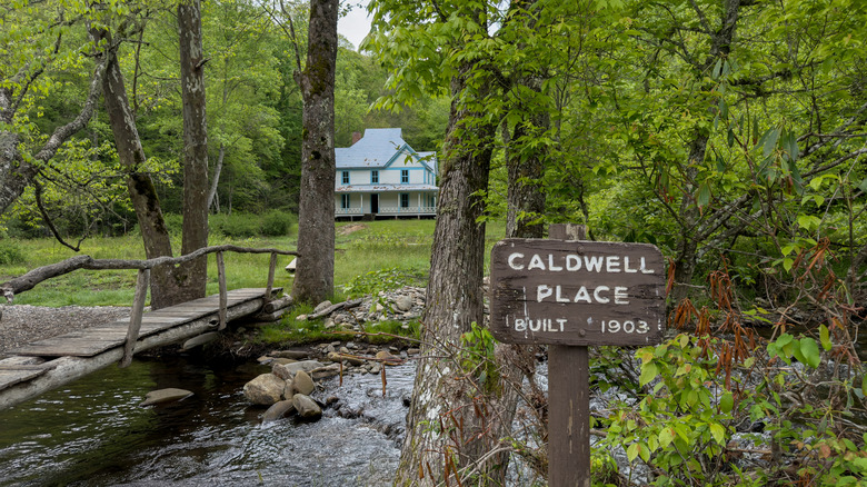 A sign introducing the 1903-built Caldwell Place and the home behind trees and a creek in Cataloochee NC