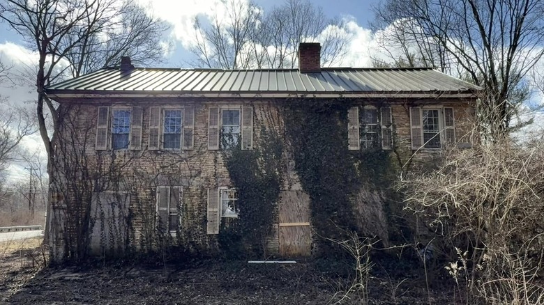 An abandoned boarding house of Carpenter's Mil -- a tan brick building with ivy growing up its front and boarded-over doorways, surrounded by winter trees under a cloudy blue sky
