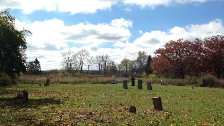 Old graves dotting a leaf-strewn field, surrounded by autumn trees under a cloudy blue sky