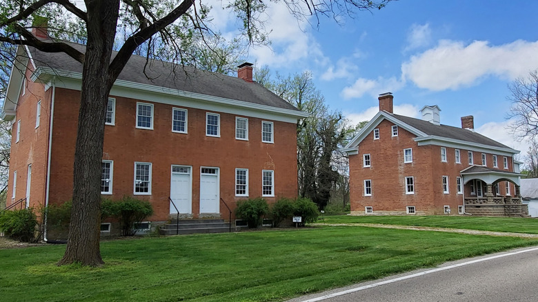 Two red brick, gray-roofed buildings, one with a stone front porch, with a large tree and green grass on front, under a cloudy blue sky