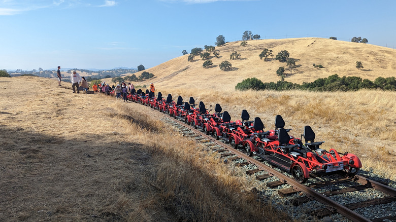 People preparing to board empty red rail bikes against tree-dotted hills of Ione, California