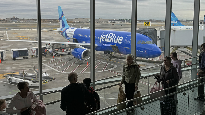 Passengers line up for boarding a plane at JFK airport with a blue JetBlue aircraft in the background out the windows