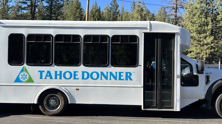 Side view of a white shuttle bus with Tahoe Donner logo parked near a wooded area on a clear day
