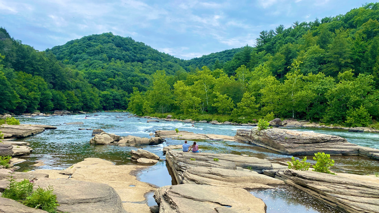 Two people in blue and pink shirts sitting on a large tan rock in a river, surrounded by more large tan rocks, with treed riverbanks and a treed mountain rising in the background, under a cloudy blue sky