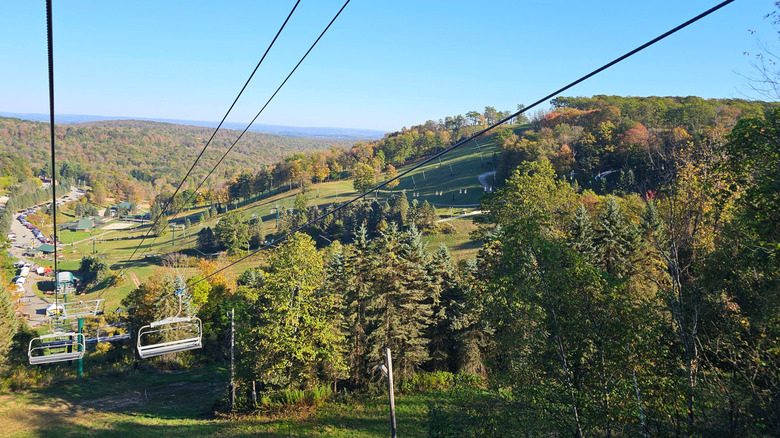 Green hills covered in trees with a mix of green and fall leaves, with a ski lift in the foreground, a parking lot along the left edge in center frame, and distant mountains in the background, under a blue sky