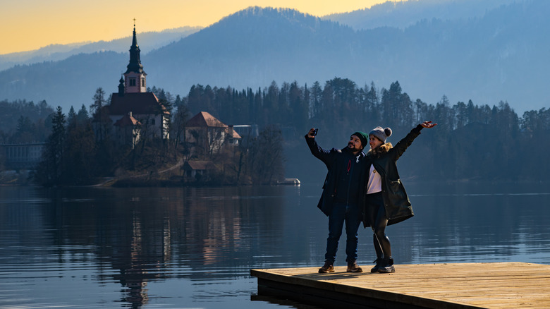 Couple takes a selfie standing on edge of a pier, lake, mountains and island in the backdrop