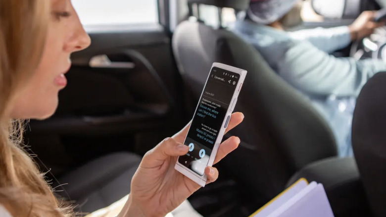 A woman seated in the back of a car uses a Vasco digital translator to speak with her driver