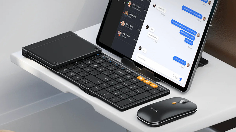 A tablet sits on a stand on an airline fold-down table with a half-folded mobile keyboard and a mouse