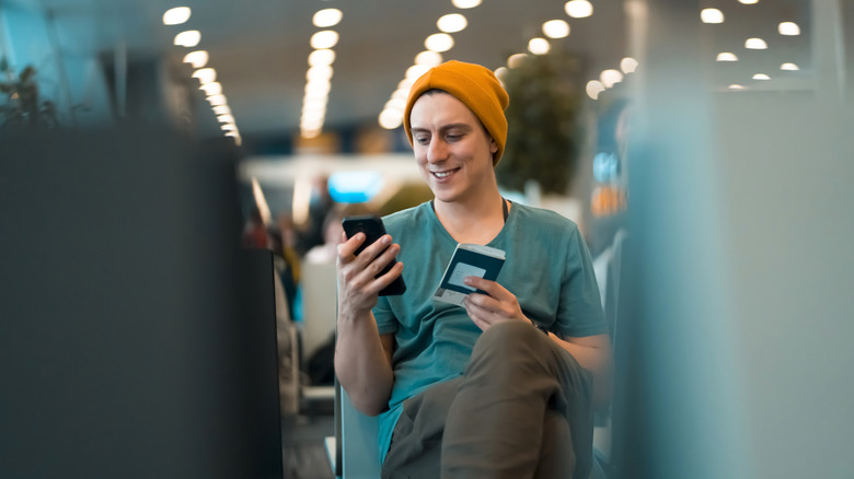 Smiling Gen Z man uses his cell phone at the airport gate to check insurance, boarding passes, and other mobile necessities