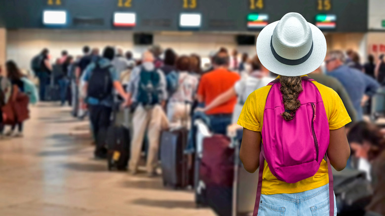 Tourist girl wearing Panama hat, yellow t-shirt, and pink backpack waiting in long lines to check in baggage at crowded airport counters.