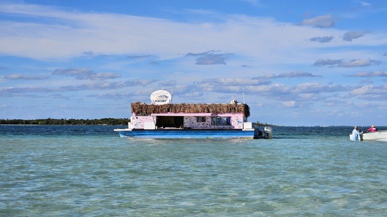 picture of the floating bar The Thirsty Cuda; it's a pink structure with a dried palm thatched roof sitting on a shallow bottomed blue boat; the bar sits in clear blue waters