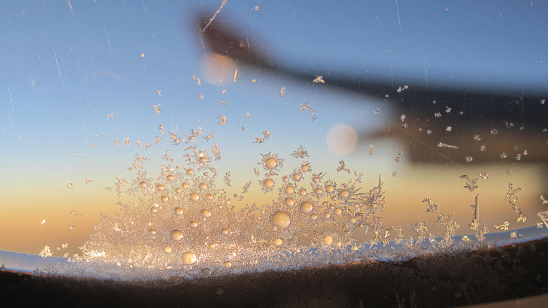 An airplane window with ice forming over it with a blurry view of an airplane wing in the background