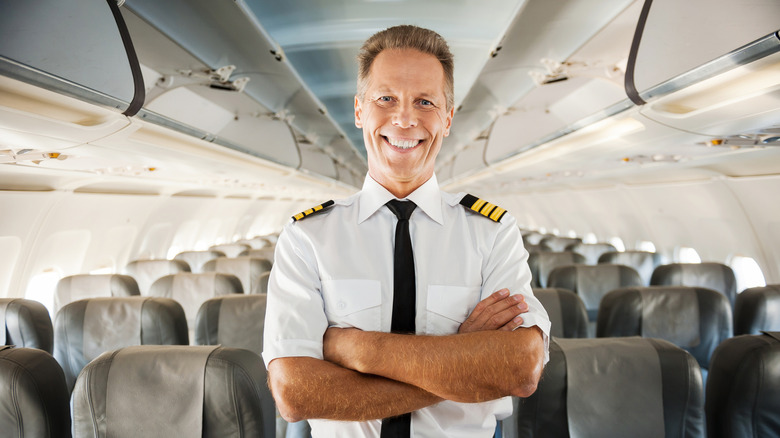 Pilot in uniform keeping arms crossed and smiling while standing inside of the airplane