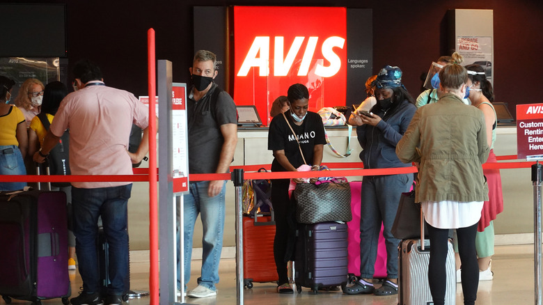 People, many wearing face masks, wait in line at Avis rental agency in the Miami International Airport Car Rental Center