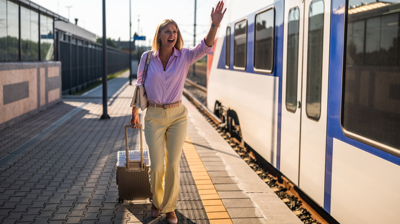 Frustrated blonde businesswoman waving to a leaving train while running along railway platform with suitcase.
