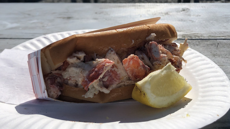 A lobster roll lying sideways on a paper plate with a slice of lemon