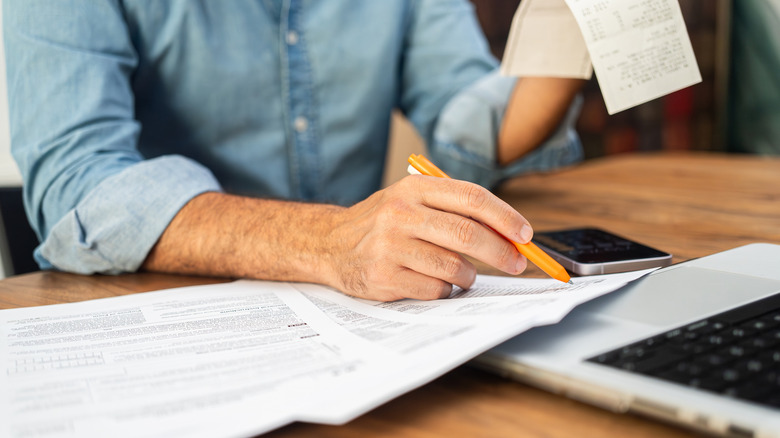 Man in a denim shirt calculating taxes with receipts and forms on a wooden desk.