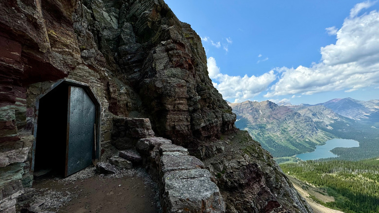 A tunnel with one metal door shut has leads through a sheer-faced mountain wall, overlooking an alpine valley in Glacier National Park