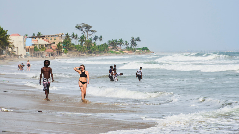 People walk on the shore and wade in the water at Kokrobite Beach