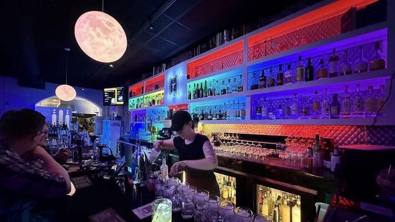 Interior of Space Bar, with a red and blue lit shelf of liquor bottles on the right, two light fixtures that look like the moon hanging from a black ceiling, and a bartender preparing a drink in the center, with two patrons sitting at the bar