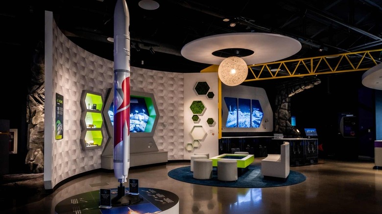 Exhibit in the Moonshot Museum, with a white and red rocket in the foreground, a curved white wall with hexagonal panels behind it, and a spherical lamp hanging over futuristic furniture, with a yellow beam and other exhibits in the background
