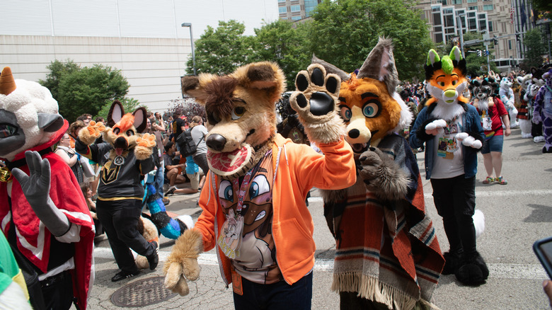 Anthrocon parade in Pittsburgh, with a brown dog fursuit carrying meat in its mouth wearing an orange hoodie in the foreground, a fox in a poncho behind them, a fox in a Vegas Golden Knights jersey to the left, and other fursuits and people around them, surrounded by buildings and trees