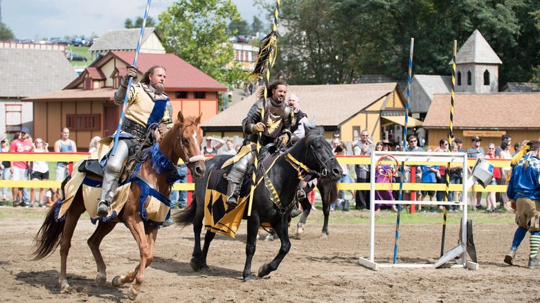 Man in yellow and blue riding a brown horse beside a man in silver armor riding a black horse with yellow and black accessories, with a crowd watching behind a yellow fence behind them and buildings and trees in the background