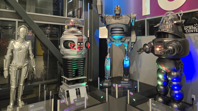 Four famous robots (Maria, B-9, the Iron Giant, and Robby) on display at the Kamin Science Center, surrounded by gray walls and windows