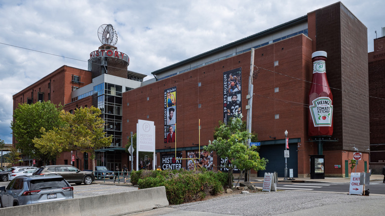 Red brick wall of the Heinz History Center with a giant ketchup pottle displayed, behind a parking lot with trees under a cloudy blue sky