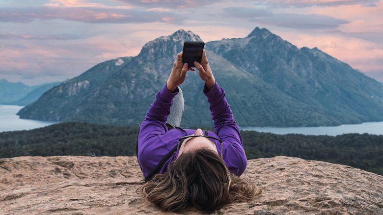 A woman looks at her phone while lying on a rock, with a large mountain and body of water in the background