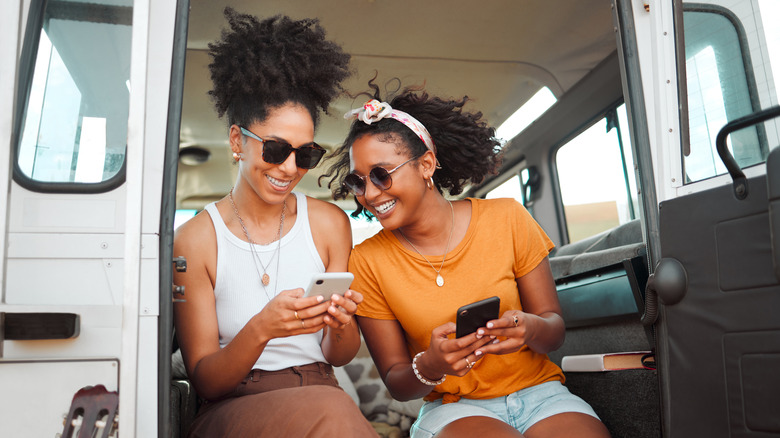 Two Black women sit inside a van laughing and looking at their phones together