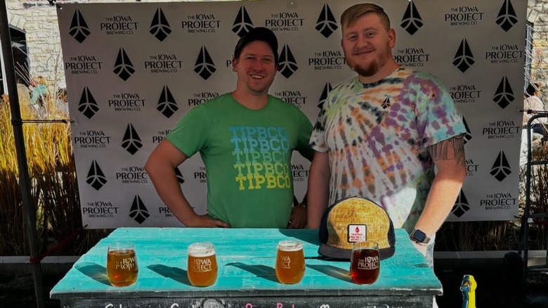 Two men stand behind a row of beer glasses, smiling
