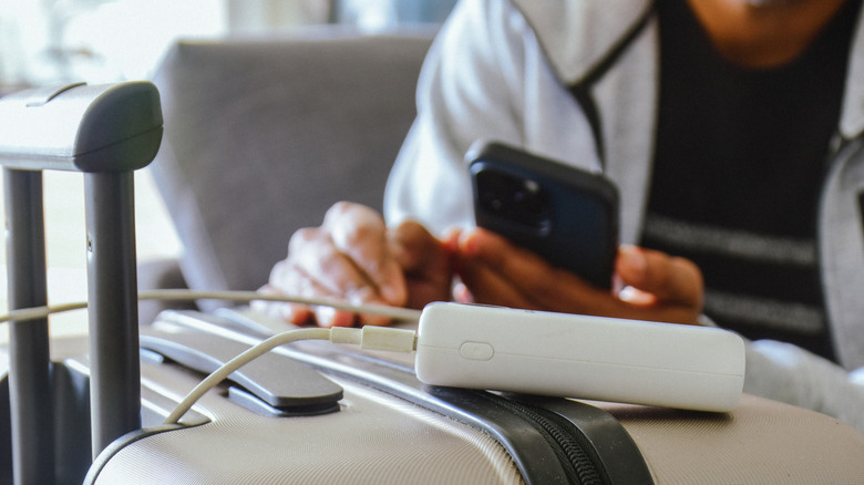 Passenger charging a black mobile phone with a white power bank and cable resting on top of a grey suitcase
