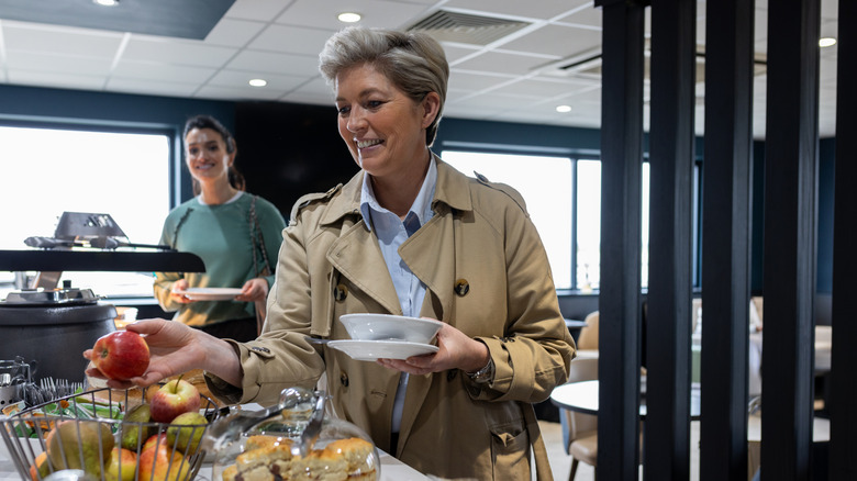 A woman in a trenchcoat taking an apple from an airport lounge buffet