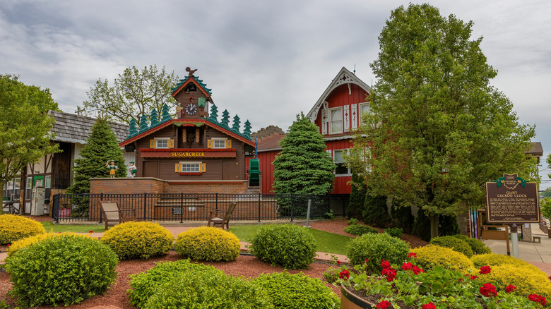 World's largest cuckoo clock in Sugarcreek, with a red building to its left behind a tree and shrubs in the foreground under a cloudy sky