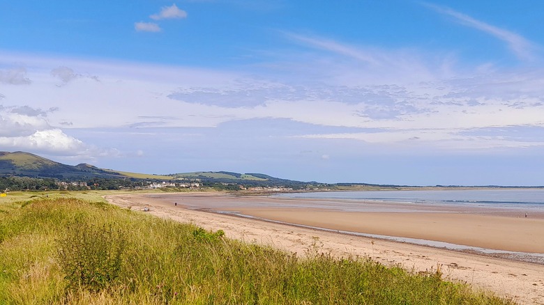 A closer view from the beach with grass in the foreground, the flat sandy beach, the sea, and low hills in the distance