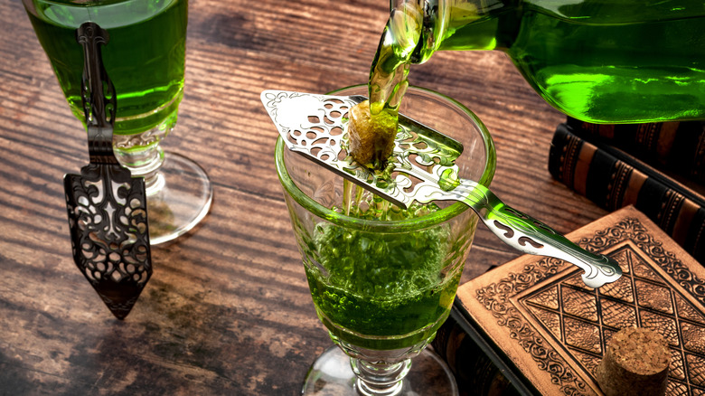 A bottle of green absinthe is poured over a special slotted spoon containing a sugar cube into a glass