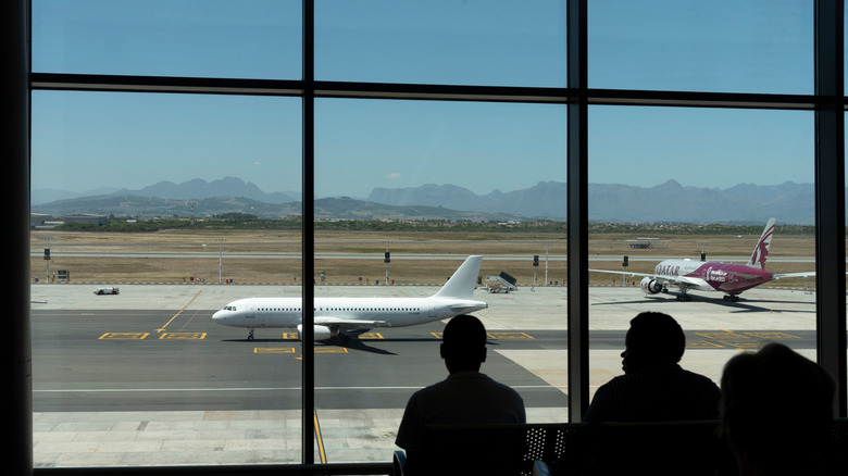 Travelers stare out window in Cape Town Airport, with mountain range visible behind the runway