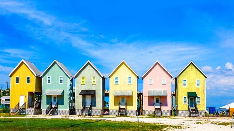 A row of colorful beach houses against a blue sky with a sandy drive and grass in front of them.