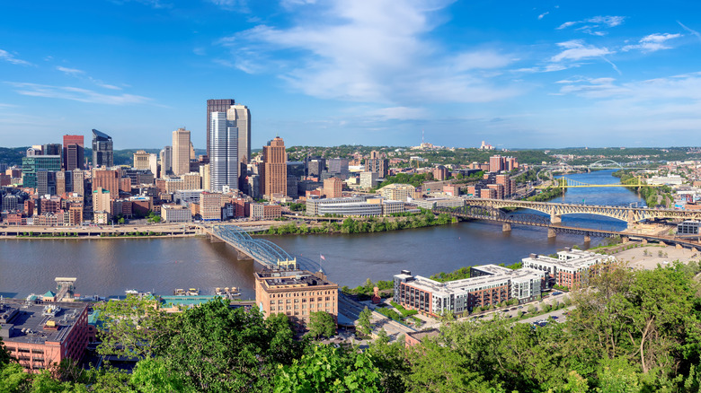 Skyline of Pittsburgh, with a river crossed by bridges and buildings surrounded by trees in the foreground, under a cloudy blue sky
