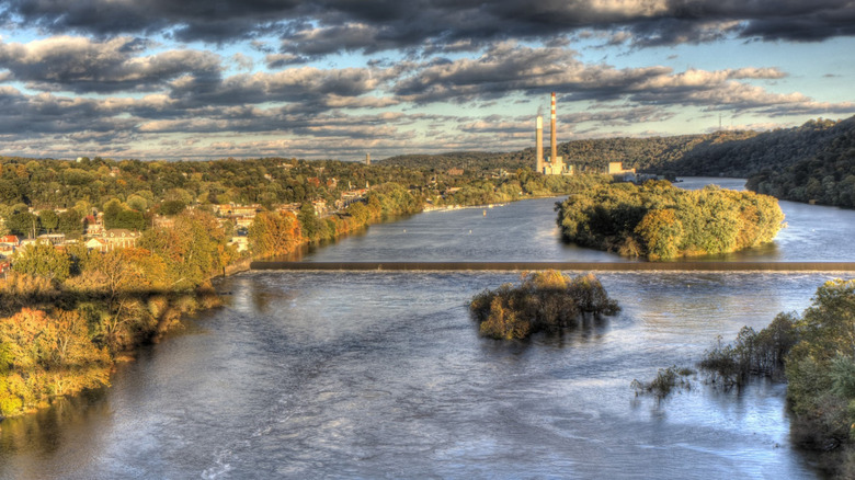 Aerial view of the Allegheny River, with a small dam separating two treed islands in center frame, a river bank with trees and buildings on the left side, and smokestacks in the background, under a cloudy blue sky