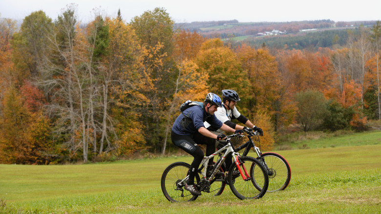 Two mountain bikers pedal across a Vermont field in front of autumnal forest.