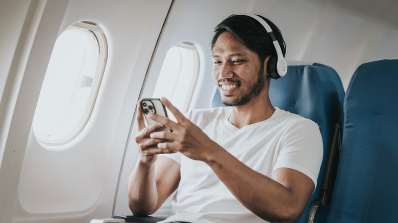 An airplane passenger wearing headphones and holding a smartphone