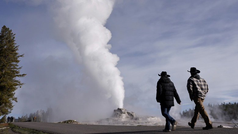 two men in cowboy hats walk towards an erupting geyser in the backdrop