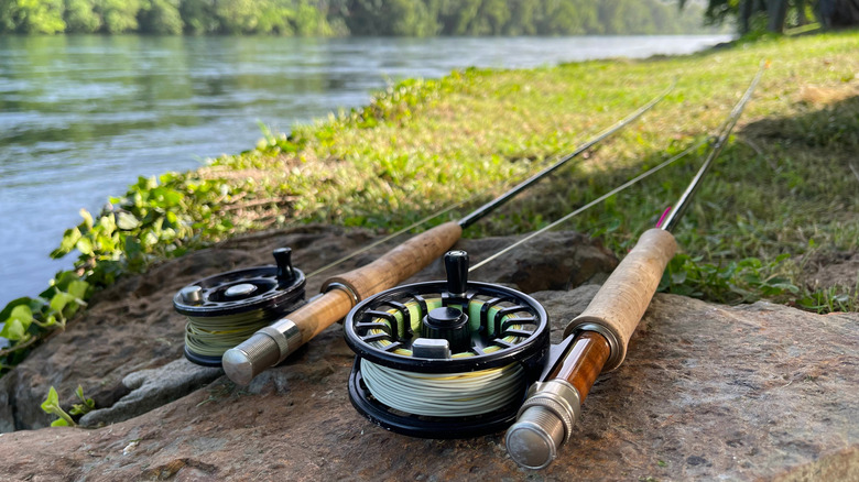 Two fly roads and reels sit on the ground next to an idyllic river