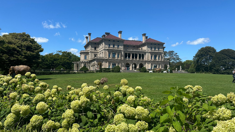 A stately mansion standing on a rolling green lawn against the blue sky, with green plantings in the foreground