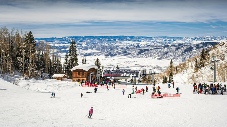 Skiers on a slope in Steamboat Springs, Colorado, with a wooden hut, ski lift, and pine trees in the mid-frame and snowy mountains in the background, under a cloudy blue sky